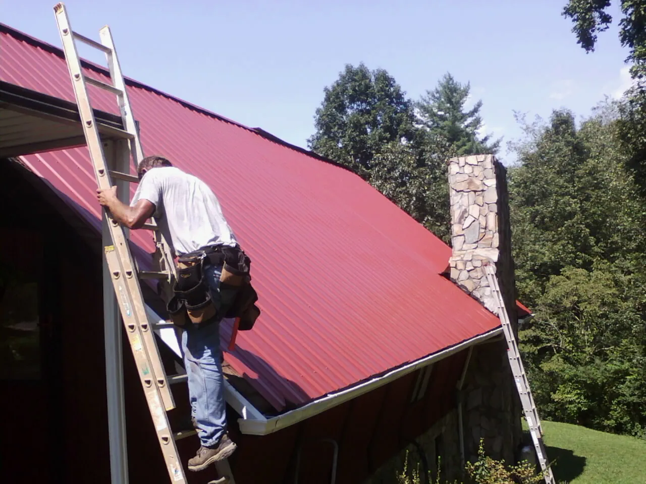 Roofer on a ladder beside a red metal roof and stone chimney — Soffit & Fascia Repair by Roofing Brothers in Catoosa